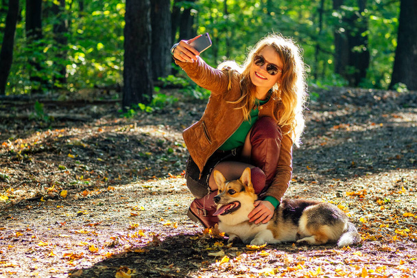 happy woman curly in a jacket and glasses mistress of a dog corgi photographing herself Selfie to the phone in the autumn park fallen yellow foliage - Valokuva, kuva