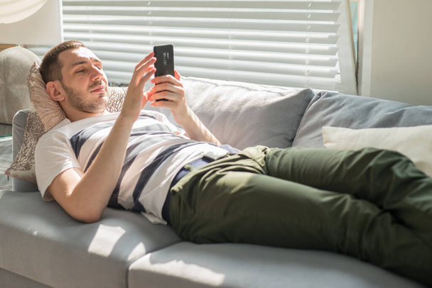 Handsome young man looking his smart phone while, lying on the couch at home - Foto, imagen