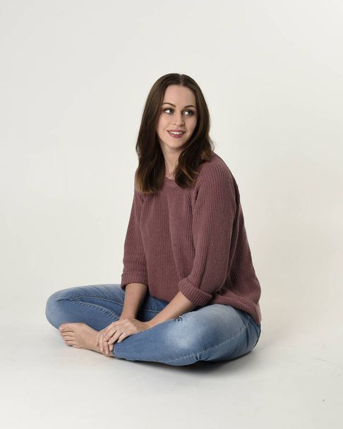 full length portrait of brunette woman wearing jeans and pink jumper. seated pose with a white studio background. - Fotografie, Obrázek