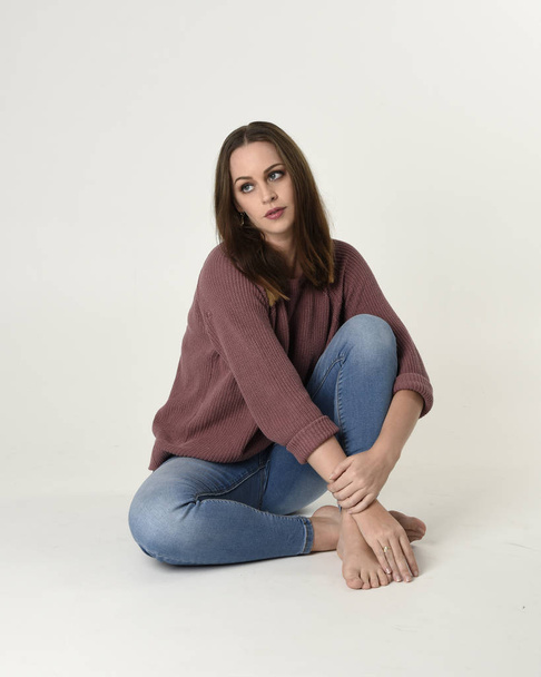 full length portrait of brunette woman wearing jeans and pink jumper. seated pose with a white studio background. - Фото, изображение