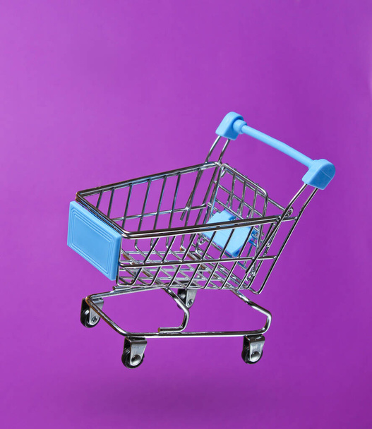 Koncepcja zakupów minimalizmu. Wózek sklepowy z zabawkami na fioletowym tle. Zdjęcie z cieniem - Zdjęcie, obraz