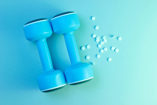 Minimalism sports pharmacology concept. Two blue plastic dumbbells and white anabolic pills, blue green holographic light. Top view - Photo, Image