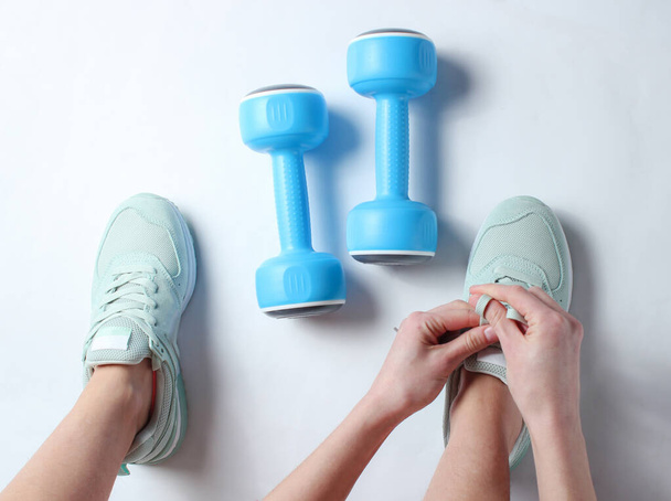 Legs of young sports woman, plastic dumbbells. Woman tying shoelace of sports shoes on a white background. Top view. Minimalism - Fotoğraf, Görsel
