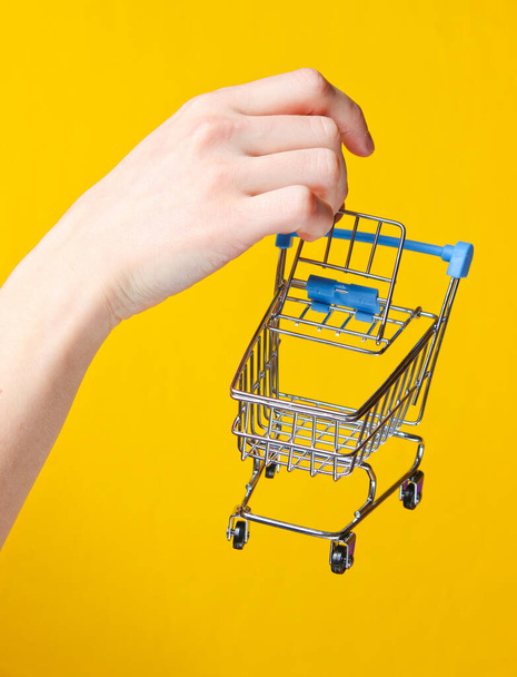 Mano femenina sosteniendo un mini carro de compras sobre fondo amarillo. Concepto de compras
 - Foto, imagen