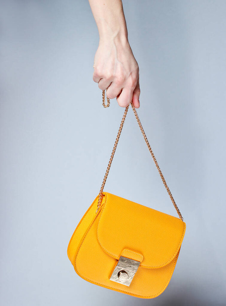 Female hand hold fashionable yellow leather bag with golden chain on gray background. - Photo, Image