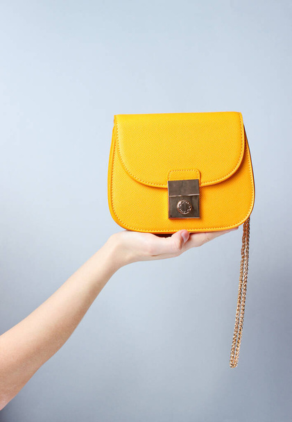 Female hand hold fashionable yellow leather bag with golden chain on gray background. - 写真・画像