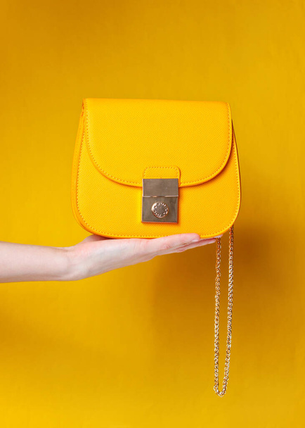 Female hand hold fashionable yellow leather bag with golden chain on yellow background. - Фото, зображення