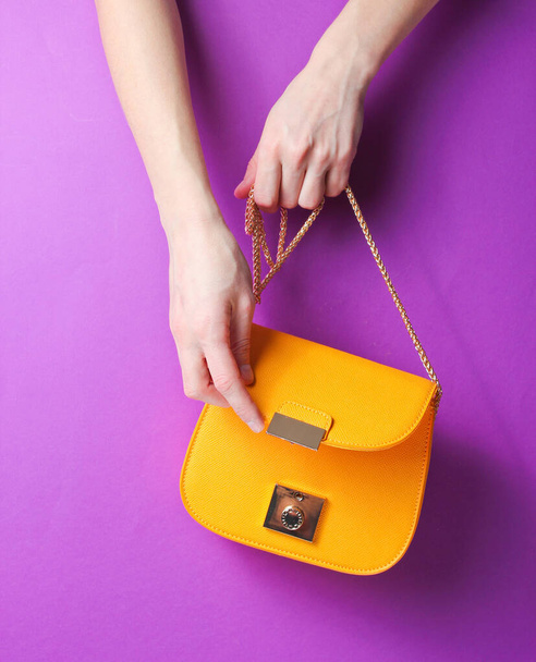 Female hand hold and opens fashionable yellow leather bag with golden chain on purple background. - Photo, Image