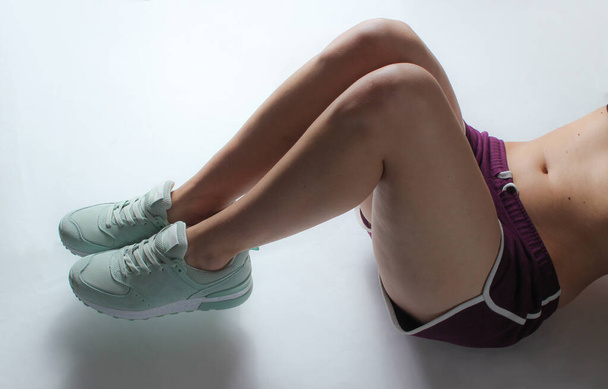Sports female body in shorts and sneakers doing double twisting exercise lying on white background - Photo, Image