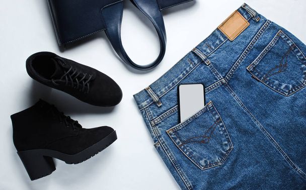 Draufsicht trendige Kleidung, Schuhe, Accessoires auf weißem Hintergrund. Jeansrock, schwarze Stiefel, Ledertasche, Smartphone in der Gesäßtasche - Foto, Bild
