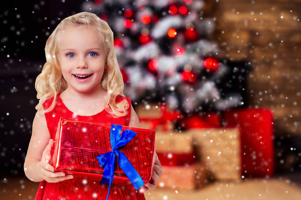 amazed surprised happy little girl blonde in red dress holding New Years gift in hand blurred christmas studio room with fireplace and new year tree.Christmas Eve morning magic and sorcery - Foto, immagini