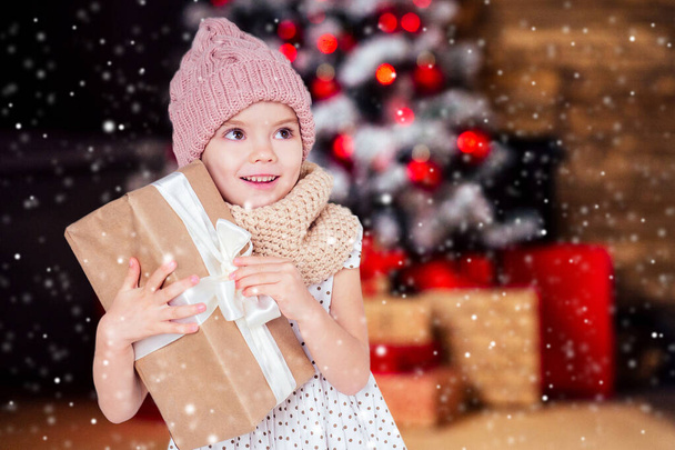 amazed surprised happy little girl blonde in white dress and pink hat holding New Years gift in hand blurred christmas studio room with fireplace and new year tree.Christmas Eve morning magic sorcery - Foto, imagen