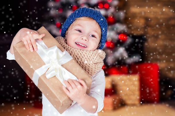 meglepett meglepett boldog kis szőke fiú fehér ing és kék kalap gazdaság szilveszteri ajándék a kezében elmosódott karácsonyi stúdió szoba kandallóval és szilveszteri fa.Szenteste reggel varázslat - Fotó, kép