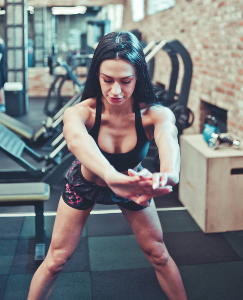 sportliche sexy brünette Frau in Sportbekleidung beim Stretching, Oberkörper nach vorne gelehnt mit den Händen vor sich selbst im Fitnessstudio - Foto, Bild