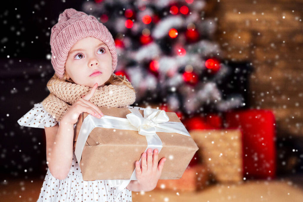 amazed surprised happy little girl blonde in white dress and pink hat holding New Years gift in hand blurred christmas studio room with fireplace and new year tree.Christmas Eve morning magic sorcery - Valokuva, kuva