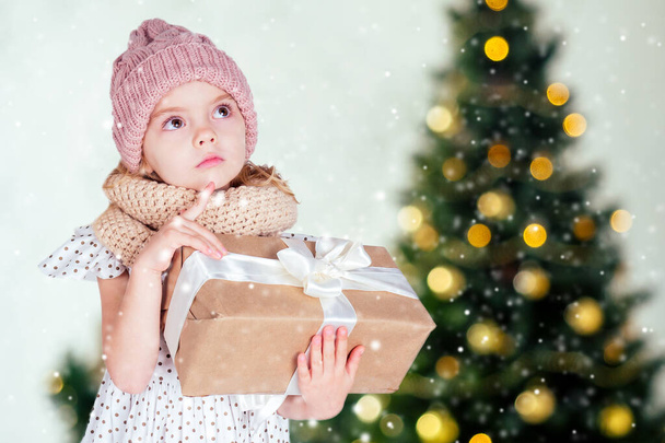 erstaunt überrascht glückliches kleines Mädchen Blondine in weißem Kleid und rosa Hut hält Neujahrsgeschenk in der Hand verschwommen Weihnachten Studio Zimmer mit Kamin und Silvesterbaum. Heiligabend Morgen Zauberei - Foto, Bild