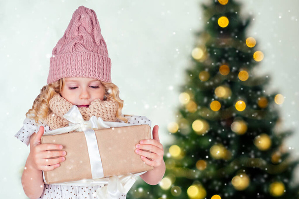 meglepett meglepett boldog kislány szőke fehér ruhában és rózsaszín kalap gazdaság szilveszteri ajándék a kezében elmosódott karácsonyi stúdió szoba kandallóval és szilveszteri fa.Szenteste reggel varázslat - Fotó, kép