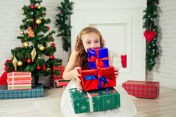 Cute little girl in a nice dress with long curly blond hair dreaming and holding a lot of gifts box at home near a Christmas tree with gifts and garlands and a decorated fireplace .new Year morning - Photo, image