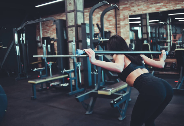 Λεπτή μελαχρινή αθλήτρια που κάνει γυμναστική με μπάρα στους ώμους της να γέρνει το σώμα της στο γυμναστήριο. Πίσω όψη - Φωτογραφία, εικόνα