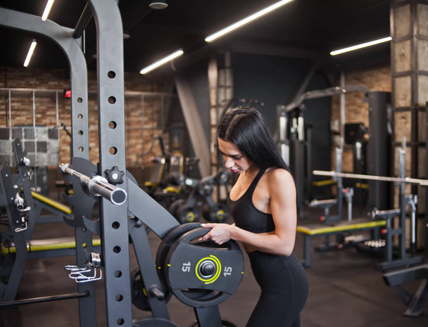Sporty brunette woman putting weight disk for barbell  against gym background - Φωτογραφία, εικόνα