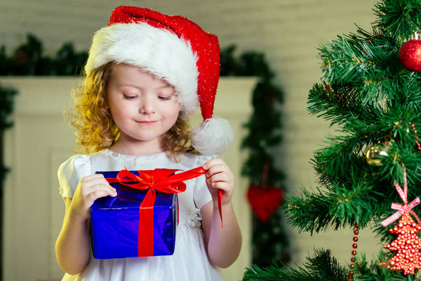 Close-up portrait of a blonde curls little girl in santa claus hat with a gift dreaming about christmas miracles standing next to the christmas tree and fireplace bright interior of the house - Фото, зображення