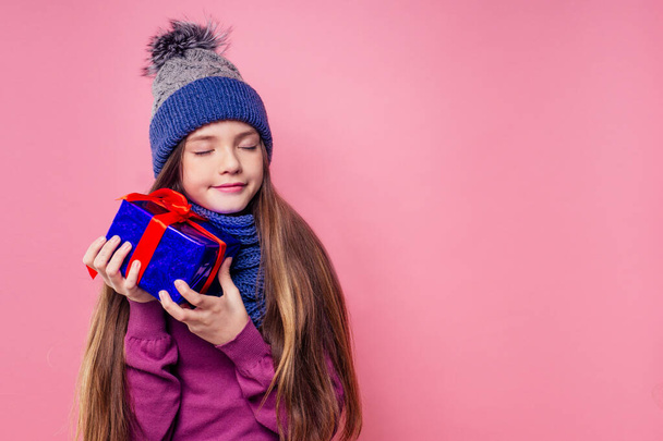 Happy little smiling blonde curly hairstyle girl in knitted blue gray hat and scarf with christmas gift box pink background studio. new Year present in hands of a female child making a wish copyspace - 写真・画像