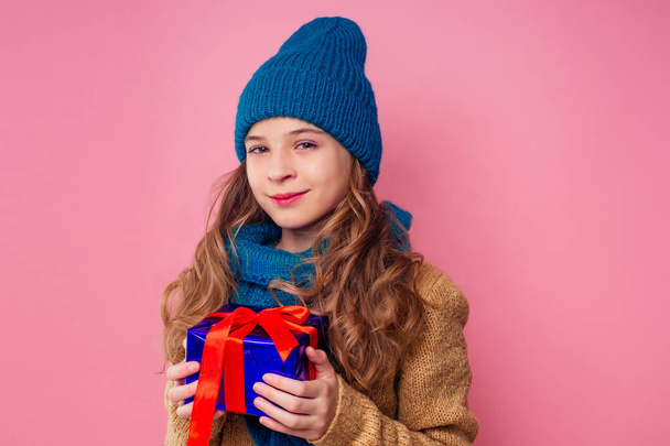 Happy little smiling blonde curly hairstyle girl in knitted green hat with christmas gift box with bow on pink background studio. new Year present in hands of a female child making a wish copyspace - Fotó, kép