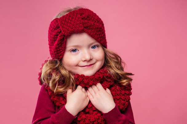 Beautiful little girl in winter warm colorful red hat and scarf dreaming thought about the gift Christmas night in the studio on a pink background.portrait photography child model - Zdjęcie, obraz