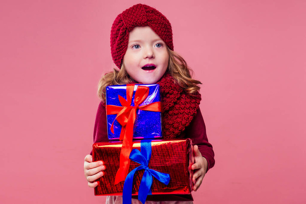 Happy little smiling blonde curly hairstyle girl in knitted gray hat with christmas gift box with bow on pink background studio. new Year present in hands of a female child making a wish copyspace - Photo, Image