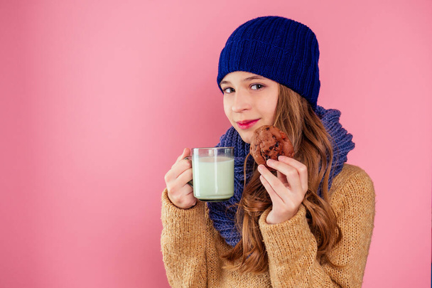 beautiful little girl drinks milk and eats cookies in a knitted hat and scarf on a pink background in the studio,christmas night - Photo, Image