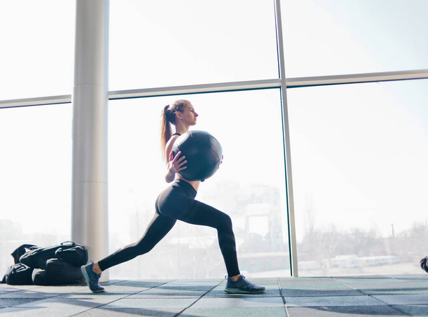 Mujer joven fitness entrenando con balón de medicina en sus manos contra el fondo de una gran ventana panorámica en el gimnasio. Entrenamiento funcional
 - Foto, imagen