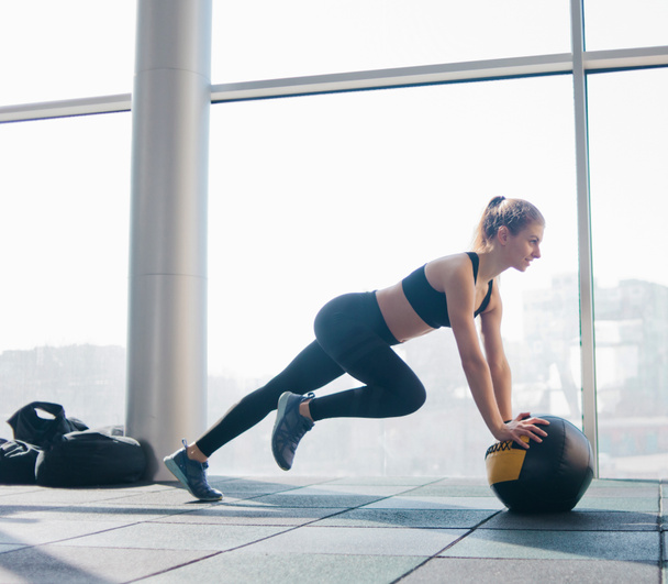 Athletic attractive woman doing exercise lifting the leg up leaning on the medicine ball against the background of large panoramic window in the gym. Functional training - Photo, Image
