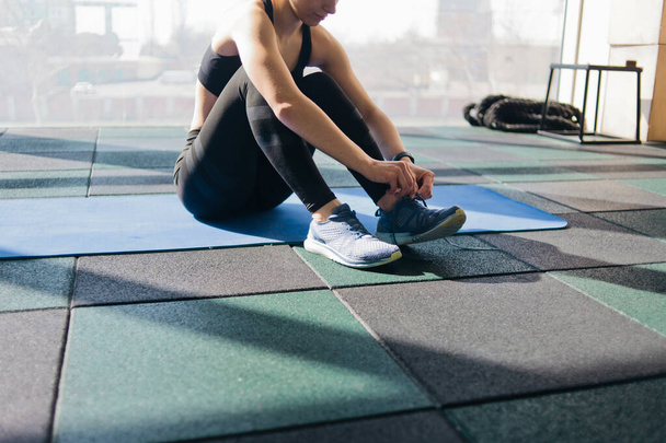 Woman tying the laces of sport sneakers sitting on a mat in the gym - Photo, Image