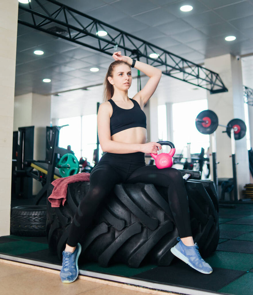 Vidám fiatal nő sportruházatban kezében vízforraló egyik kezével ül egy nagy kerék funkcionális edzés és pihenés a tornateremben. - Fotó, kép