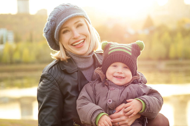 Mother in a warm jacket and knitted hat hugging her baby boy walking in the autumn park. stylish blondewoman with a child against the backdrop of the lake - Φωτογραφία, εικόνα