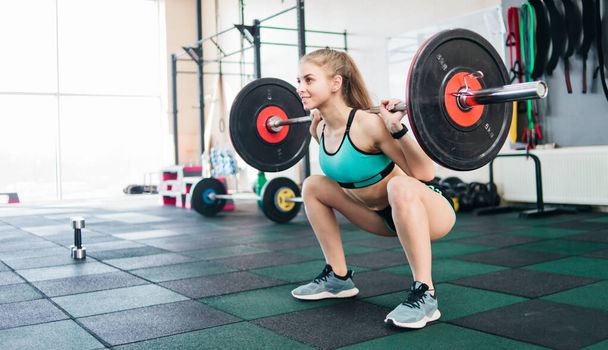 Young blonde woman doing squatting with a barbell on the shoulders in the gym - Photo, Image