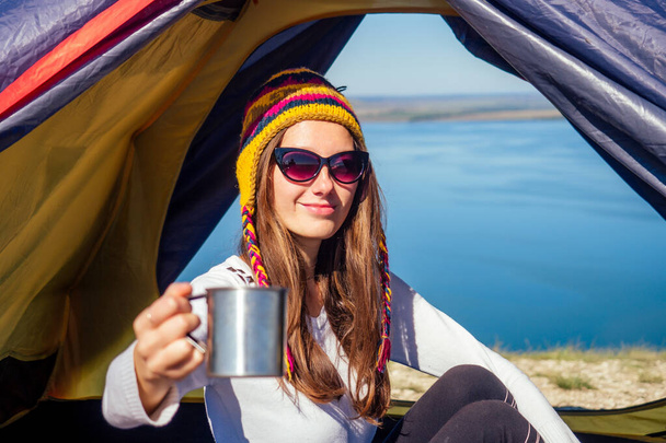 Young woman in a funny hat from Nepal sitting in a blue sleeping bag in the tent holding a thermos mug and drinking coffee cup tea background landscape lake. good morning in the journey - Fotografie, Obrázek