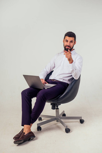 confident and handsome male office worker smiling and holding a laptop sitting on a chair on a white background in the studio full-length portrait - Photo, image