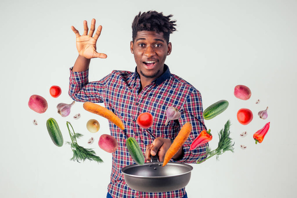 african american chef kitchener holding a frying pan wizard man cooking magic flying food salad, carrot, garlic, onion, pepper, potato, cucumber isolate white background studio.magic of taste fantasy - Fotografie, Obrázek