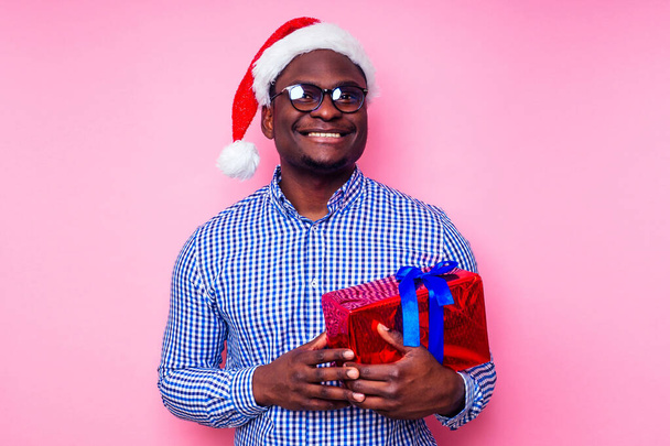 Afro-Amerikan adam şık ekose gömlek giyiyor Noel Baba şapkalı, pembe arka plan stüdyosunda hediye kutusu olan harika bir gülümseme. Koyu tenli Noel Baba mutlu noeller kutluyor. - Fotoğraf, Görsel