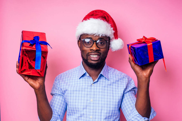 African American man wearing stylish plaid shirt great smile in santa hat with gift box on pink background studio.dark-skinned Santa Claus congratulating merry christmas - Photo, image