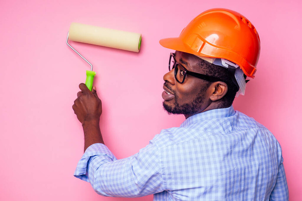 black man African American holding paint roller in hand paints the wall in pink color .happy african builder painting inside the house,businessman wears a helmet hard hat.young guy is painting - Foto, afbeelding