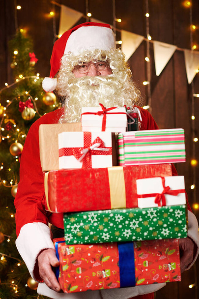 Άγιος Βασίλης ποζάρουν με πολλά δώρα, κάθονται σε εσωτερικούς χώρους κοντά διακοσμημένα χριστουγεννιάτικο δέντρο με φώτα - Καλά Χριστούγεννα και καλές διακοπές! - Φωτογραφία, εικόνα