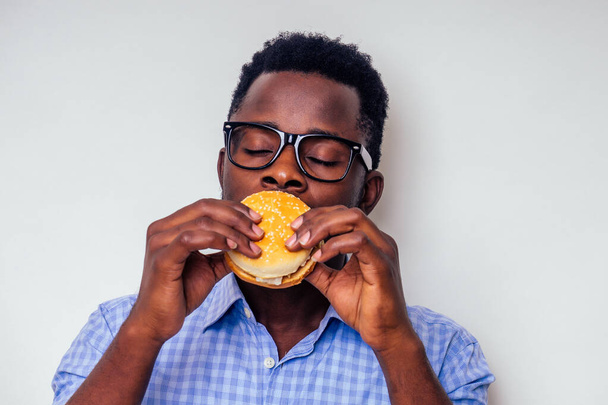african american man enjoying the taste of hamburger.handsome and young afro man in a stylish shirt and glasses holding a burger on a white background.love of junk food diet - Foto, imagen