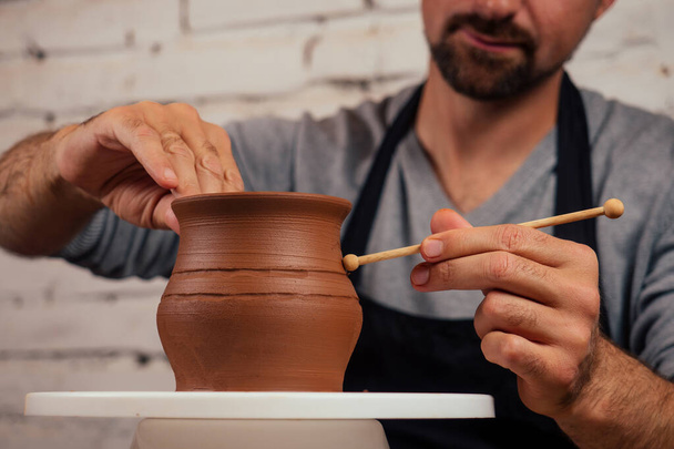 handsome young potter man in a gray sweater and a black apron sculpts clay pot on a potters wheel in a cozy craft workshop. businessman owner of a dishes china shop - Foto, Imagen
