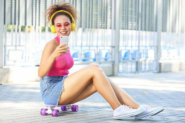 Hermosa afroamericana con monopatín y teléfono móvil escuchando música al aire libre
 - Foto, Imagen