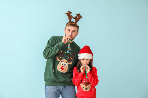 Funny little girl and her father in Christmas sweaters and with party whistles on color background - Foto, Bild