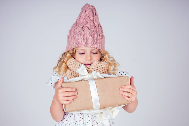 Happy little smiling blonde curly hairstyle girl in a knitted pink hat with christmas gift box with bow on white background in studio. new Year present in the hands of a female child making a wish - 写真・画像