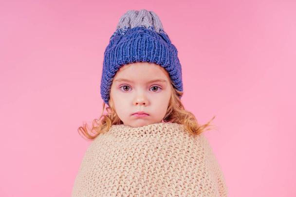 portrait of unhappy little girl wearing knitted scarf and sweater and scarf. Child getting sick flu common cold on pink background in studio. viral autumn winter season chill quarantine concept. - Foto, Imagem
