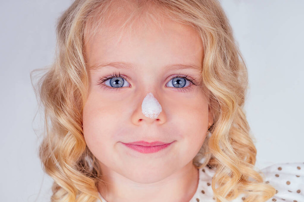 little and beautiful blonde curly hairstyle girl in cute dress blot sunscreen on the face. blue eyes, perfect skin, on the nose spf cream close up portrait on white background in studio - Foto, afbeelding